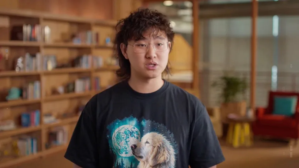 A person standing indoors in front of bookshelves, wearing a graphic T-shirt.
