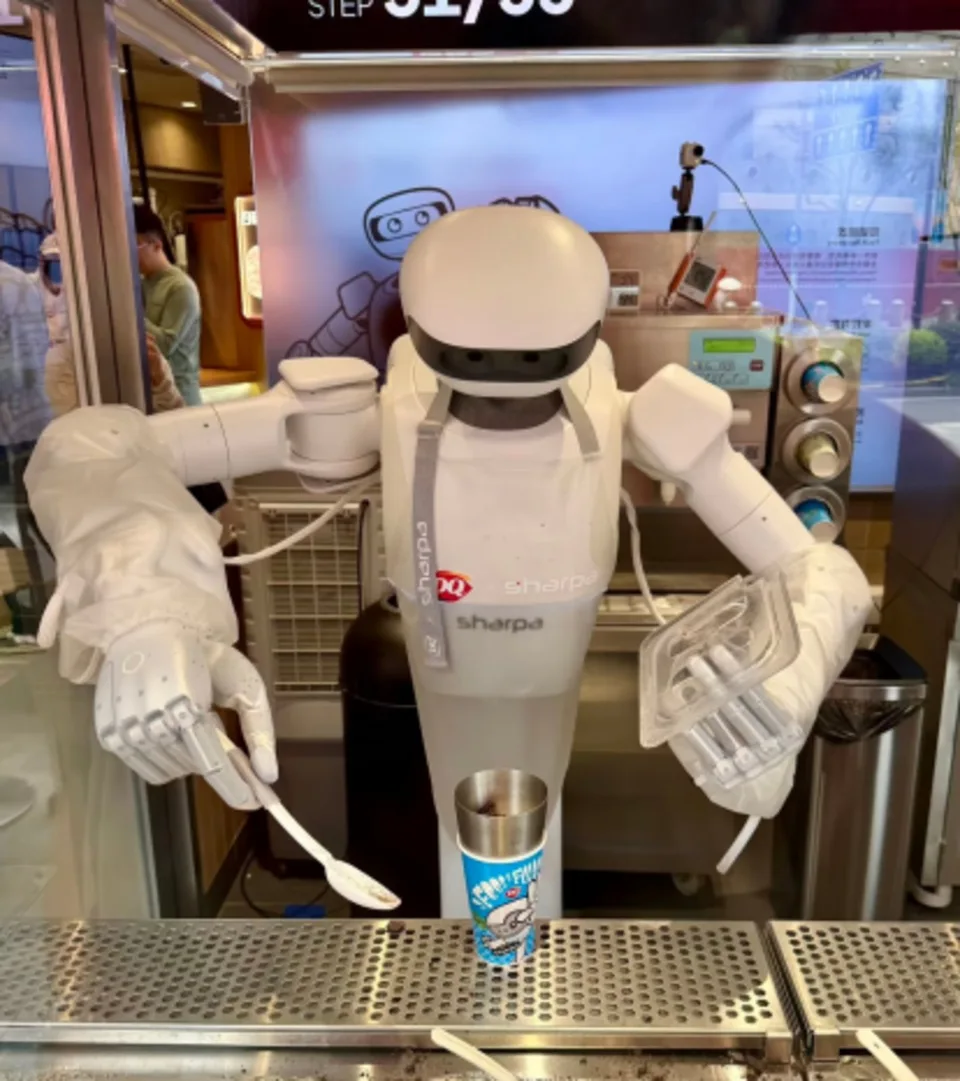 A humanoid robot stands behind an ice-cream counter holding a cup and utensil.