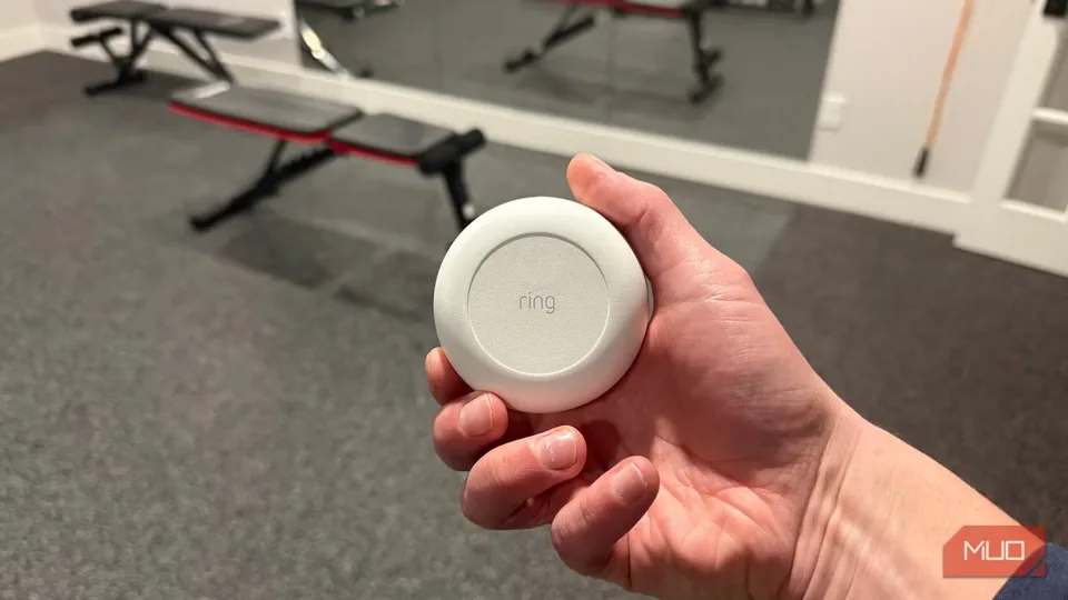 A hand holds a small round Ring sensor in an office setting.