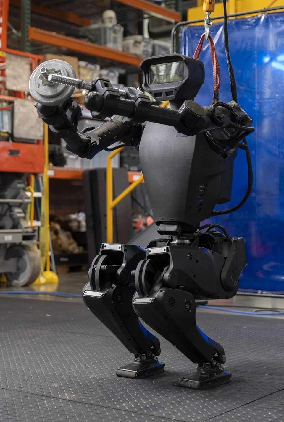A humanoid robot crouches while holding a bar-like load in an industrial test area.