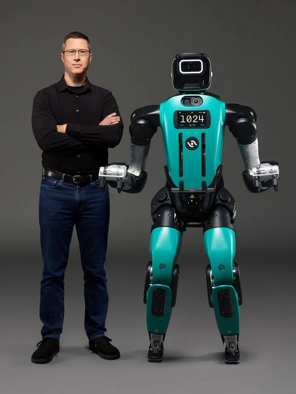 Agility Robotics’ teal humanoid Digit robot stands beside a person in a studio portrait.
