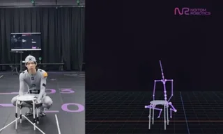 A humanoid robot seated beside a small table in a motion-capture space, with a monitor visible behind it.