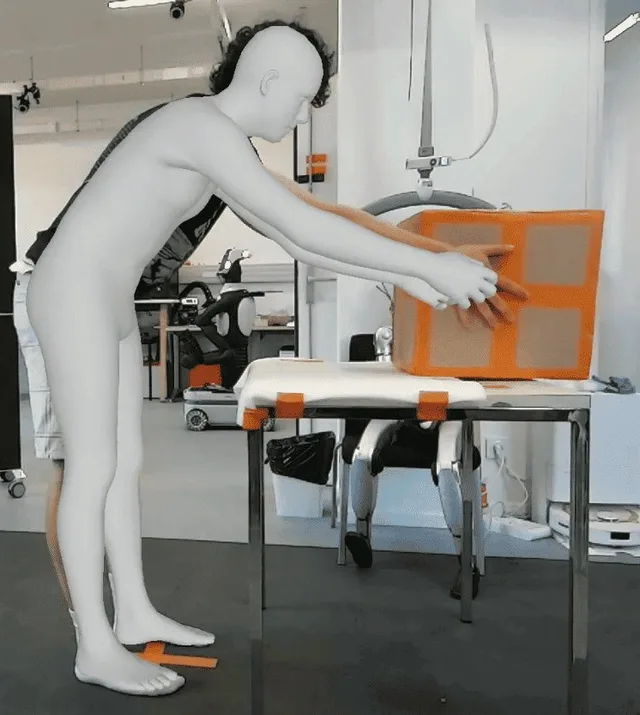 A humanoid robot reaches toward an orange box on a table in a laboratory.