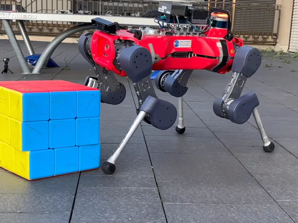A red quadruped robot stands beside a large blue-and-yellow block on an outdoor paved surface.