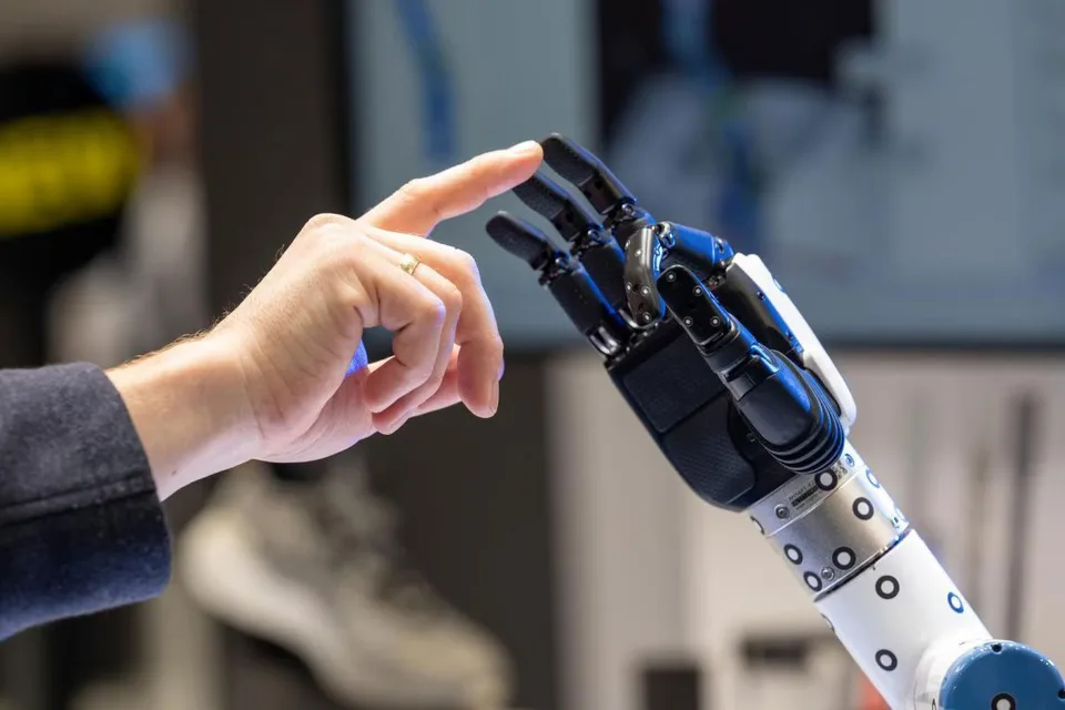 A person reaches toward a robotic hand in a factory-like setting.