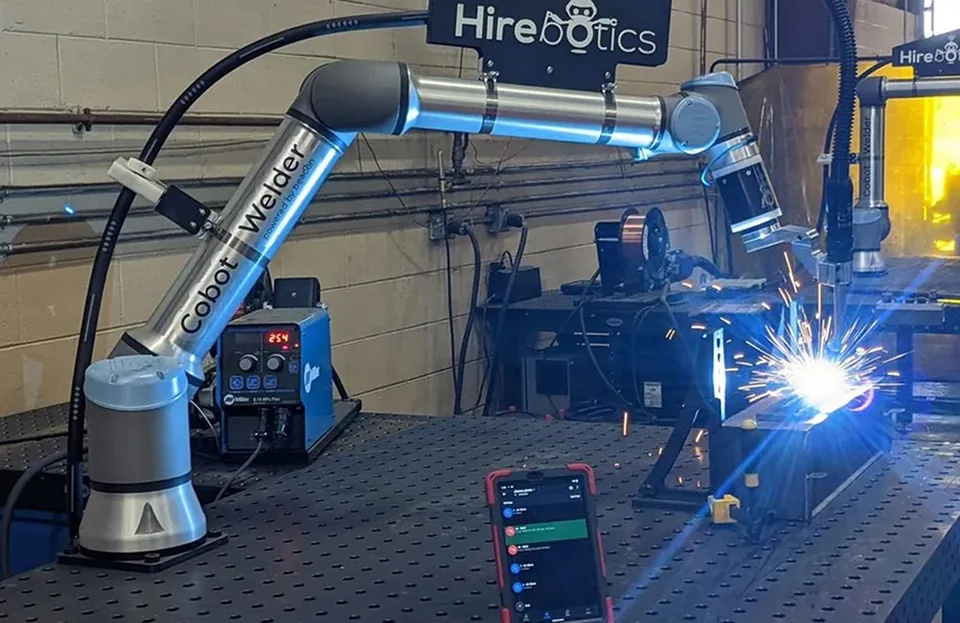 Hirebotics collaborative robot welding at a workbench beside a person using a tablet to control a cobot arm.