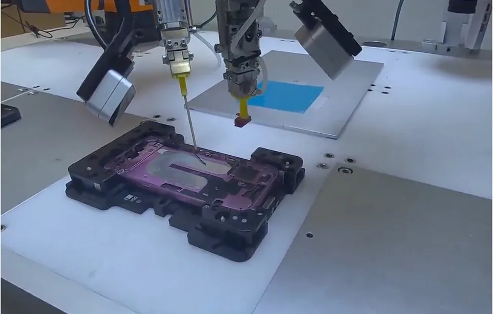 Industrial robotic arms use tools over a component on a workbench.
